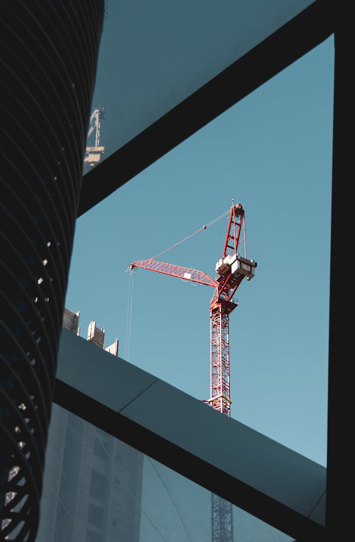A red tower crane in a city construction setting framed by modern architecture, clear blue sky.